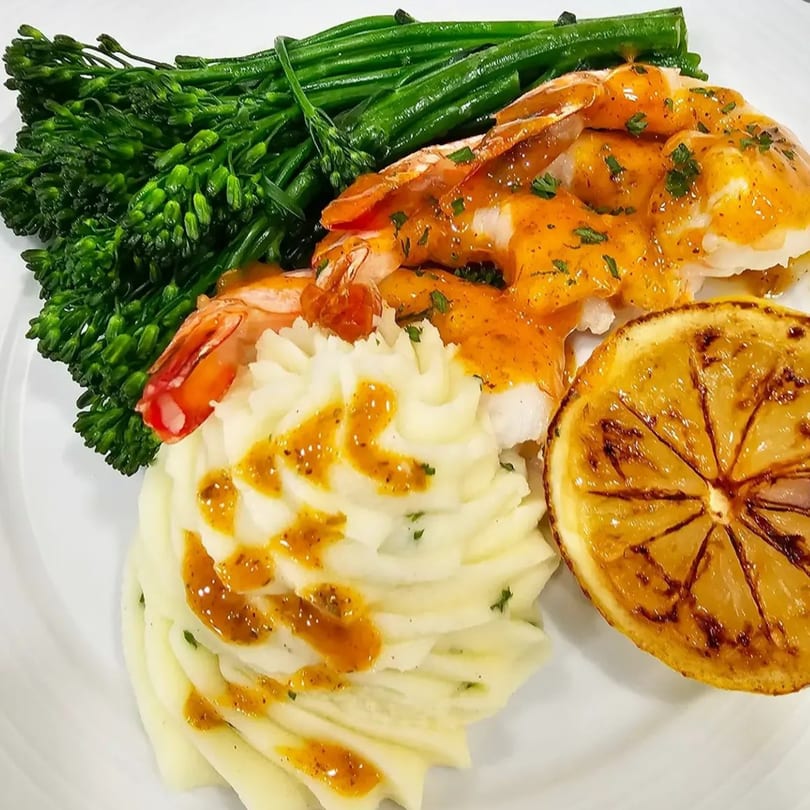 Shrimp with sauce, mashed potatoes, broccolini, and a grilled lemon on a white plate.