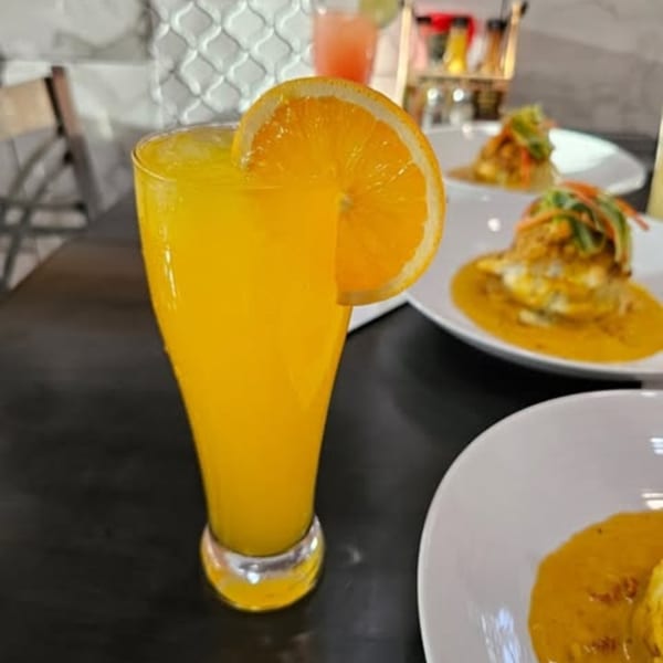 Orange juice with an orange slice garnish, and two plates of food in the background.