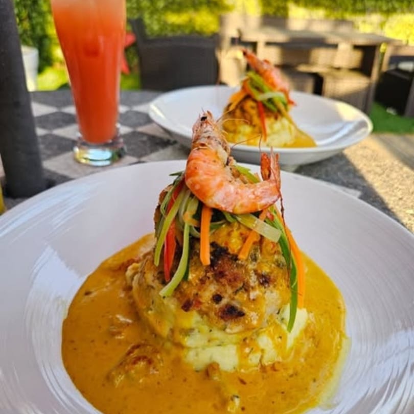 Two plates of shrimp and crab cakes with sauce, mashed potatoes, and a drink on an outdoor table.