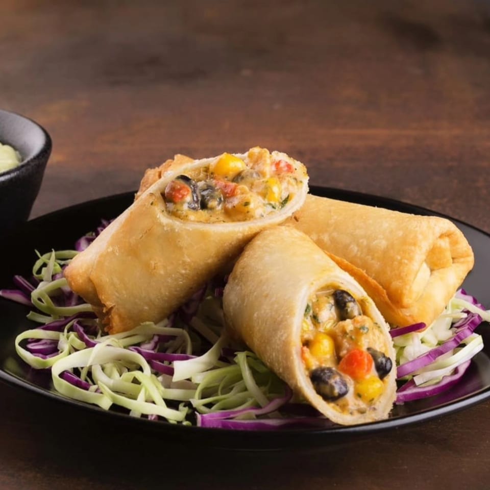 Three golden-brown egg rolls filled with corn, black beans, and red peppers, served on shredded cabbage.
