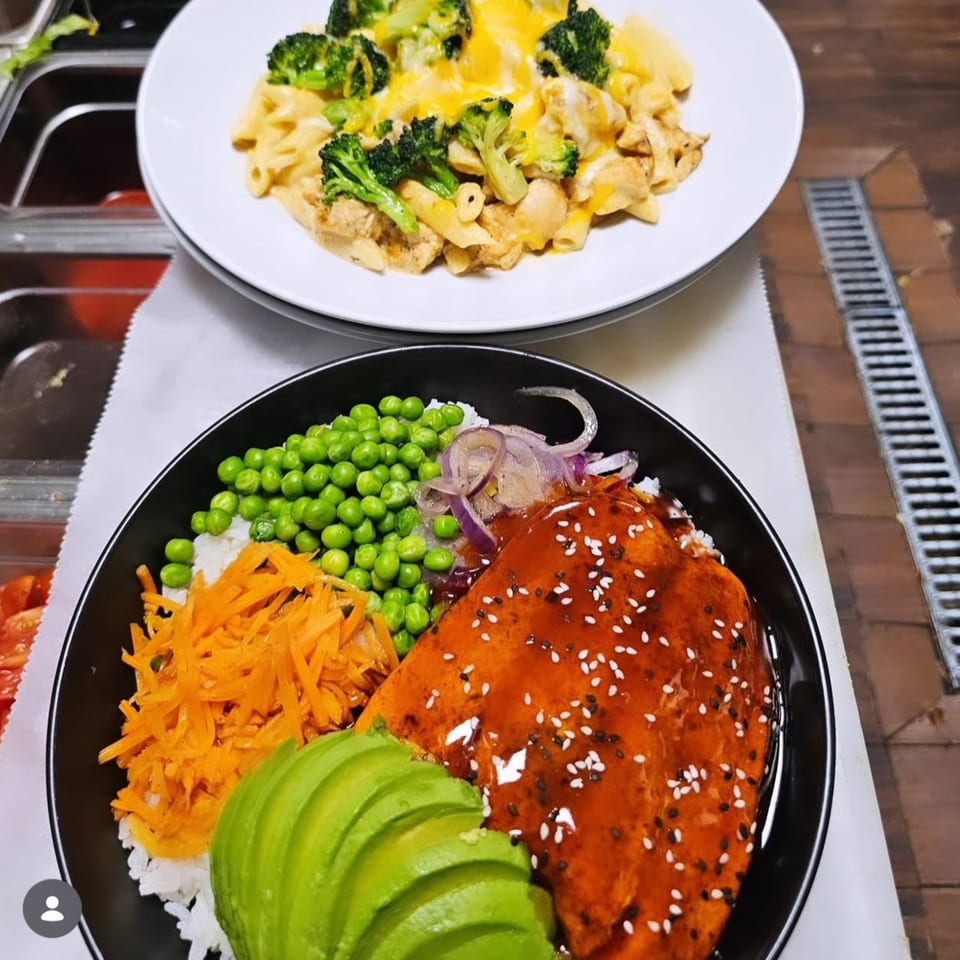 A salmon poke bowl with avocado, peas, carrots, and rice, next to a plate of macaroni and cheese.