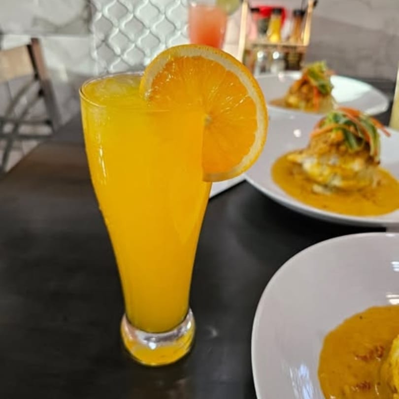 Orange juice with an orange slice garnish, next to plates of food on a dark table.