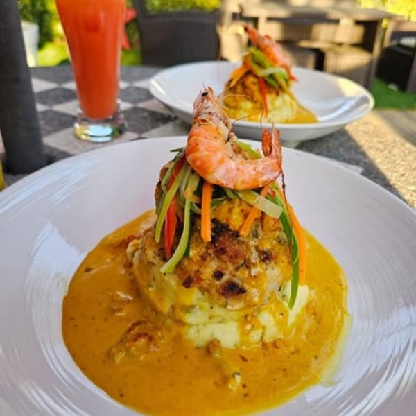 Two plates of shrimp and vegetable-topped seafood cakes with yellow sauce, and a red drink.