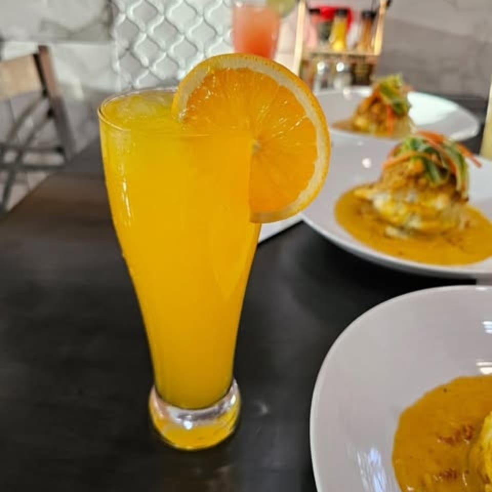 A tall glass of orange juice with an orange slice, next to plates of food.
