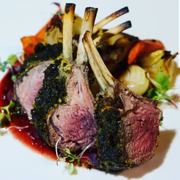 Herb-crusted lamb chops, medium-rare, with roasted vegetables and a red sauce on a white plate.